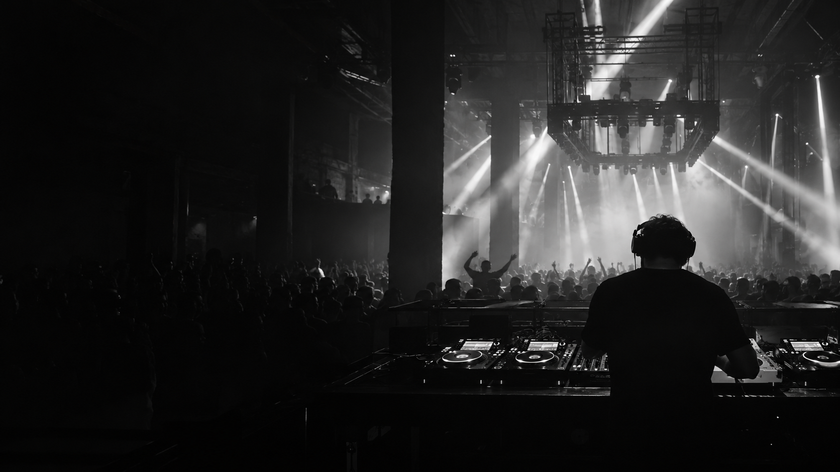 DJ at a nightclub booth, facing a crowd under white stage lights